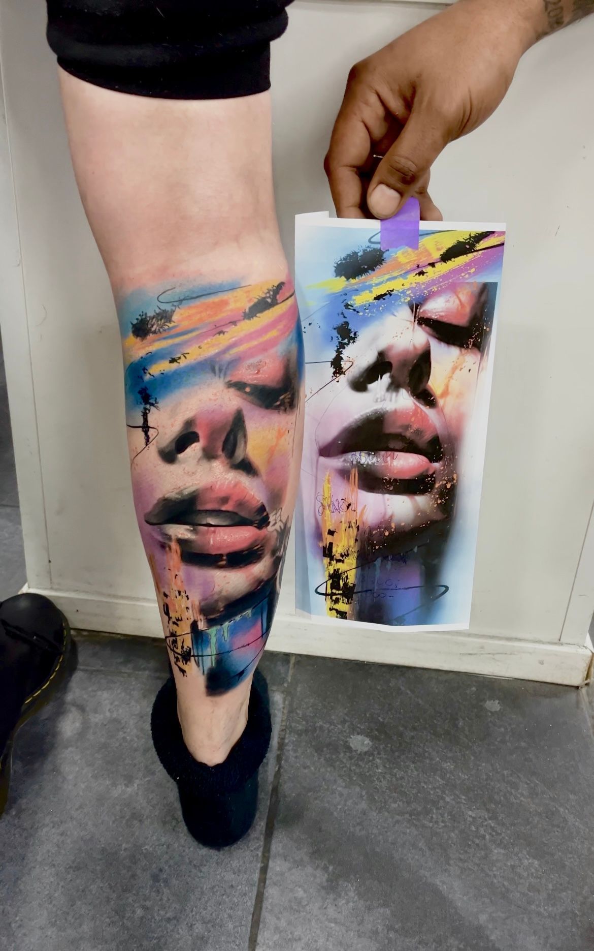 Tatoeage op een been van een kleurrijk gezicht met gesloten ogen, naast een soortgelijke print.