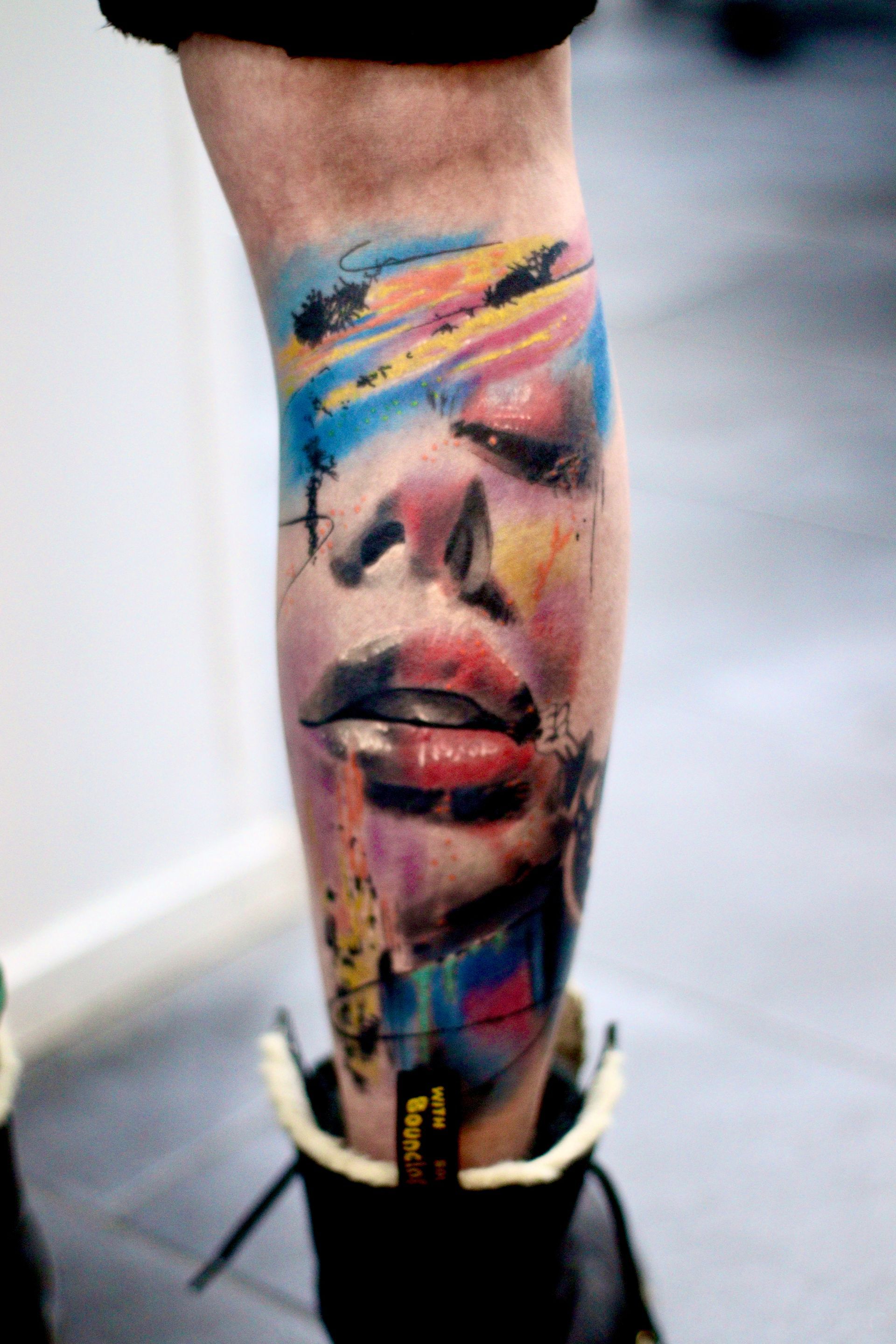 Levendige tatoeage van een gezicht met volle lippen op een been. Omringd door kleurrijke penseelstreken en spetters.