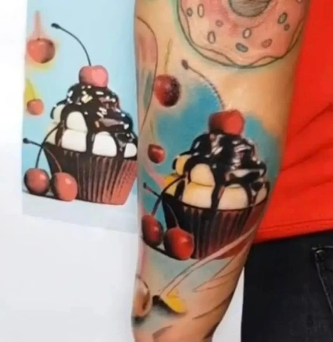 Cupcake-tatoeage op de arm, met kersen en chocolade, met blauwe en gele accenten.