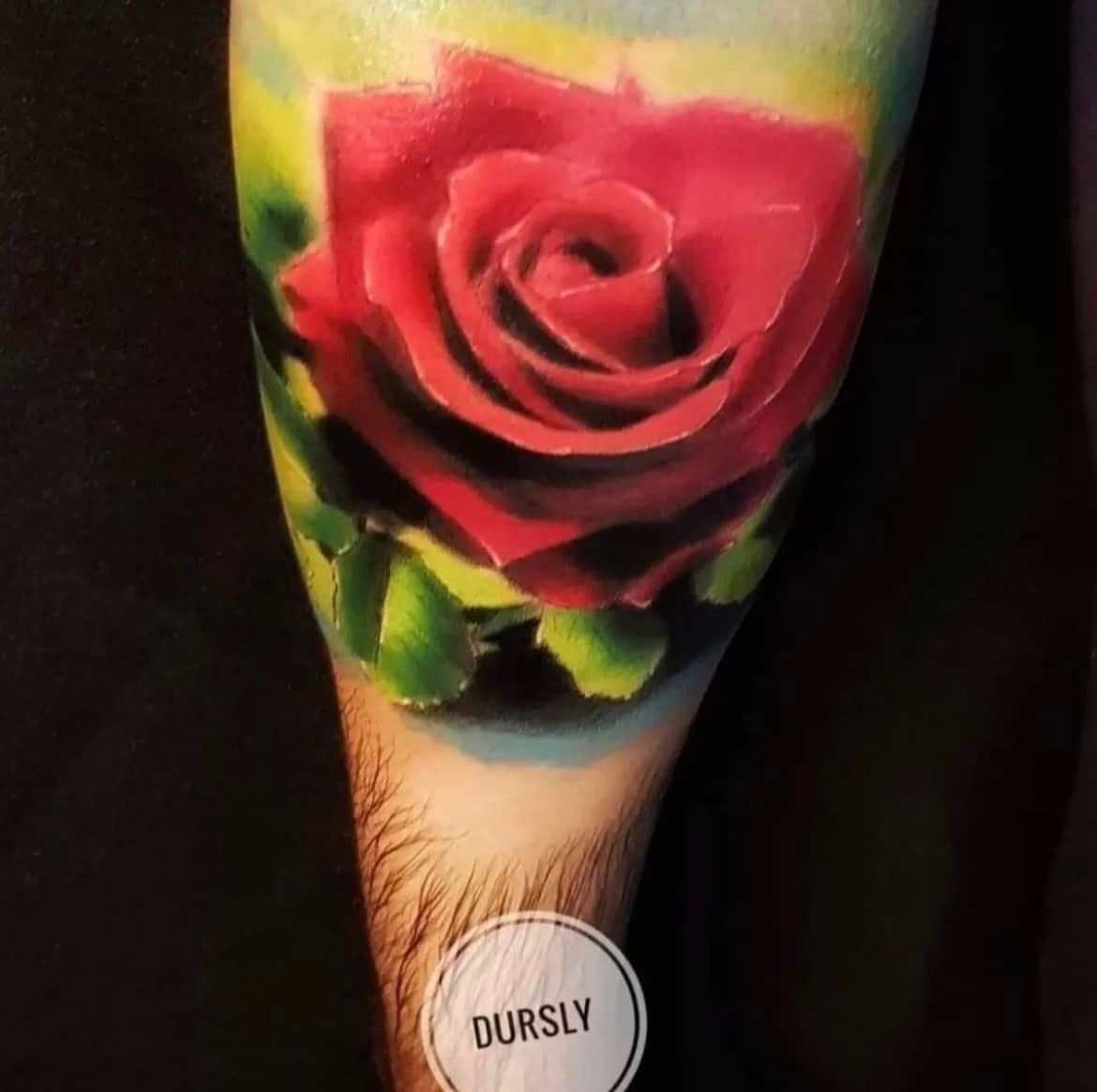 Levendige rode rozentatoeage op de arm, met groene bladeren, tegen een vage geel en blauwe achtergrond.