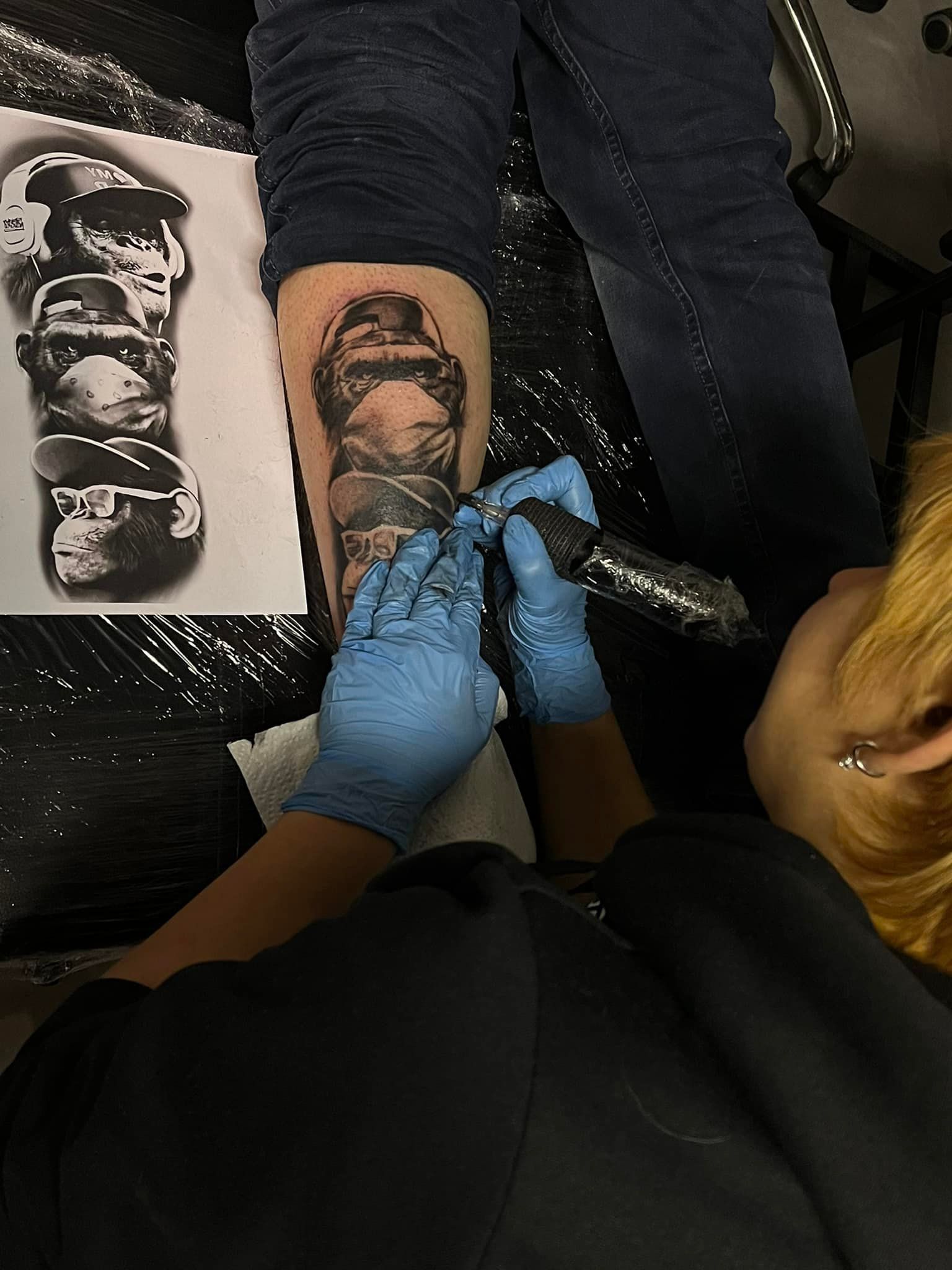 Tatoeëerder met blauwe handschoenen die een ontwerp op een been maakt. Referentiebeeld van auto's in de buurt.