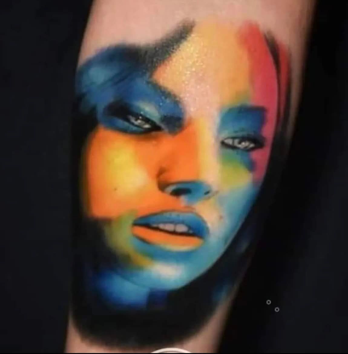 Kleurrijke tatoeage van een gezicht, voornamelijk blauw, oranje en geel. Intense blik, open mond.