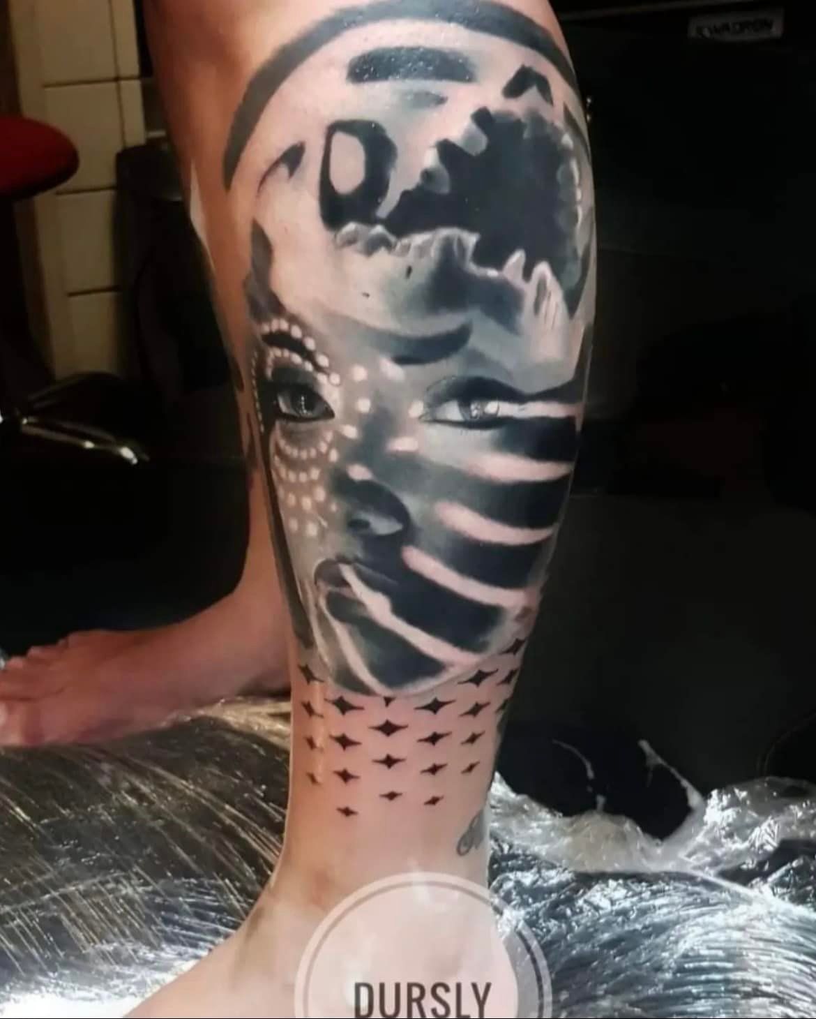 Zwart-grijze tatoeage op haar been: vrouwengezicht gecombineerd met een maan, sterren eronder, die getatoeëerd wordt.