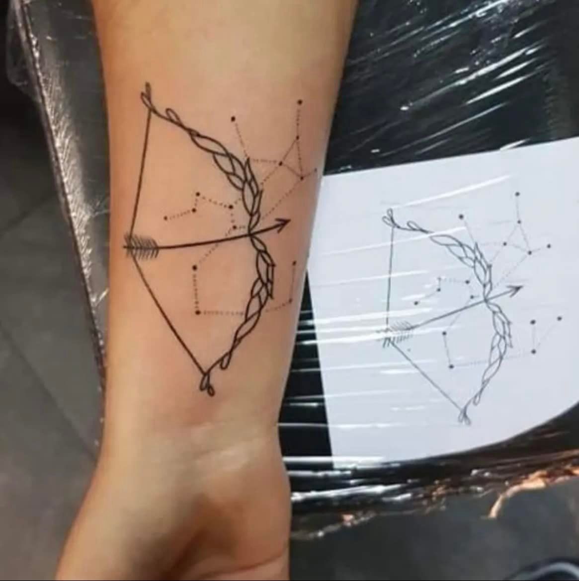 Tatoeage van een pijl en boog met daaraan verbonden sterrenbeelden op de pols van een persoon.