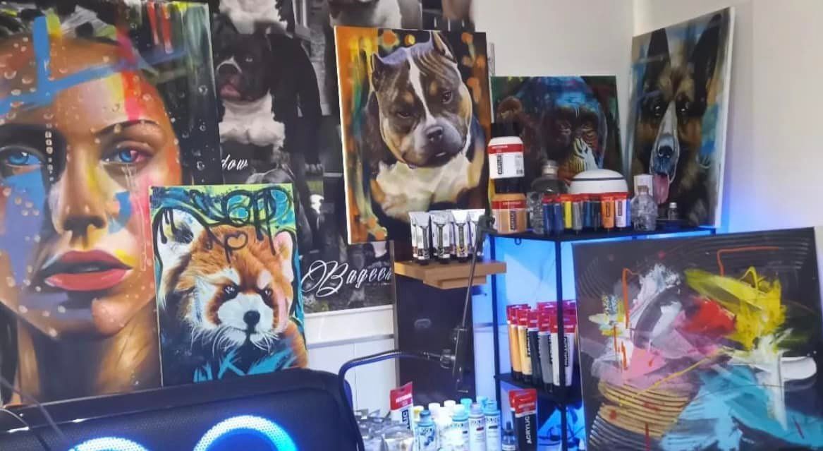 Atelier vol kunstwerken: schilderijen van een vrouw, een hond en dieren. Verftubes en spuitbussen zijn zichtbaar.