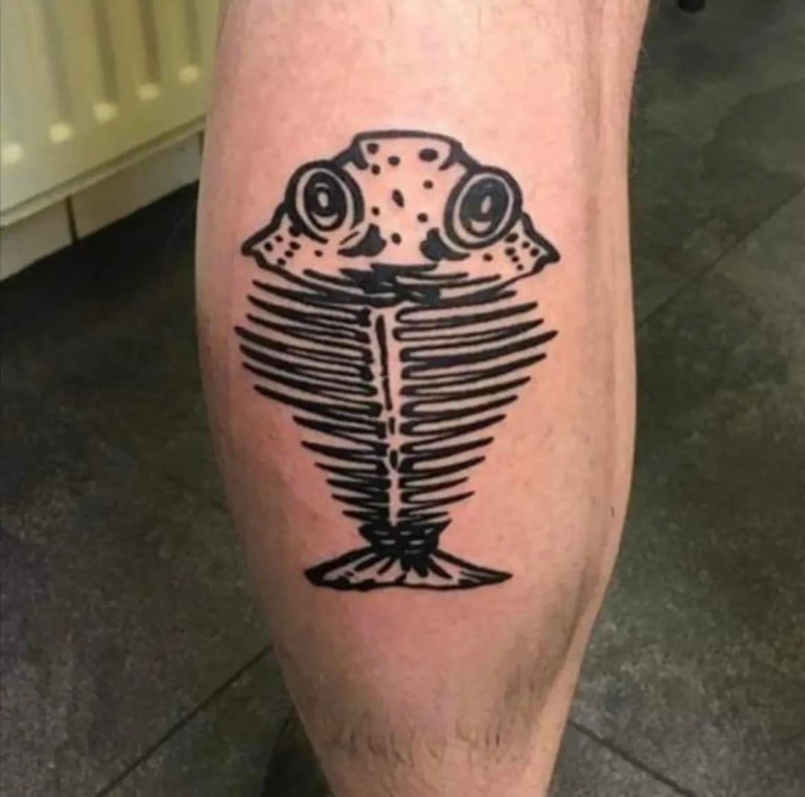 Zwarte tatoeage van een visskelet met kikkerachtige kenmerken op het been van een persoon.