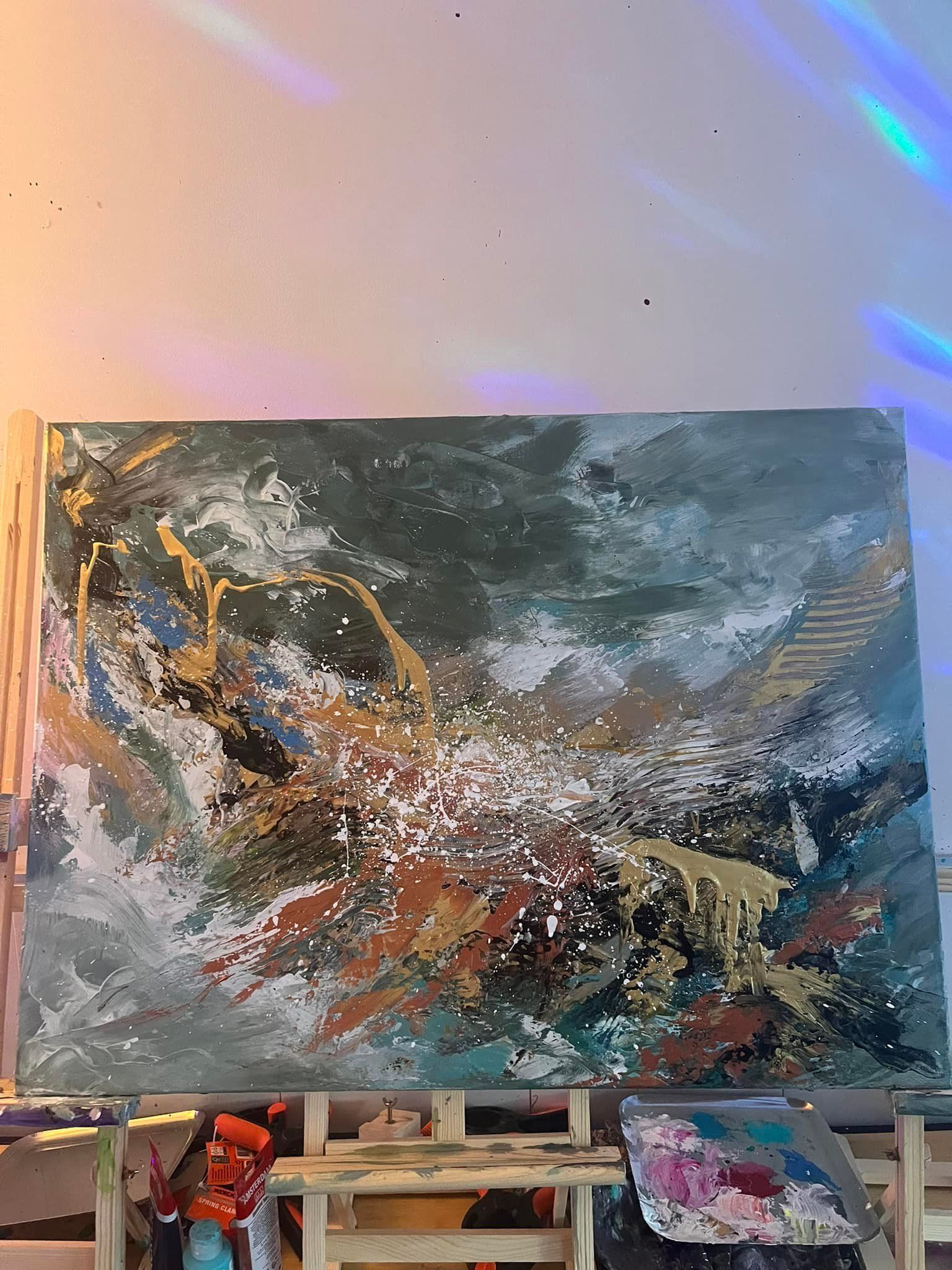 Abstract schilderij op een houten ezel. Blauwgroene, gouden en witte penseelstreken creëren een landschap met textuur.