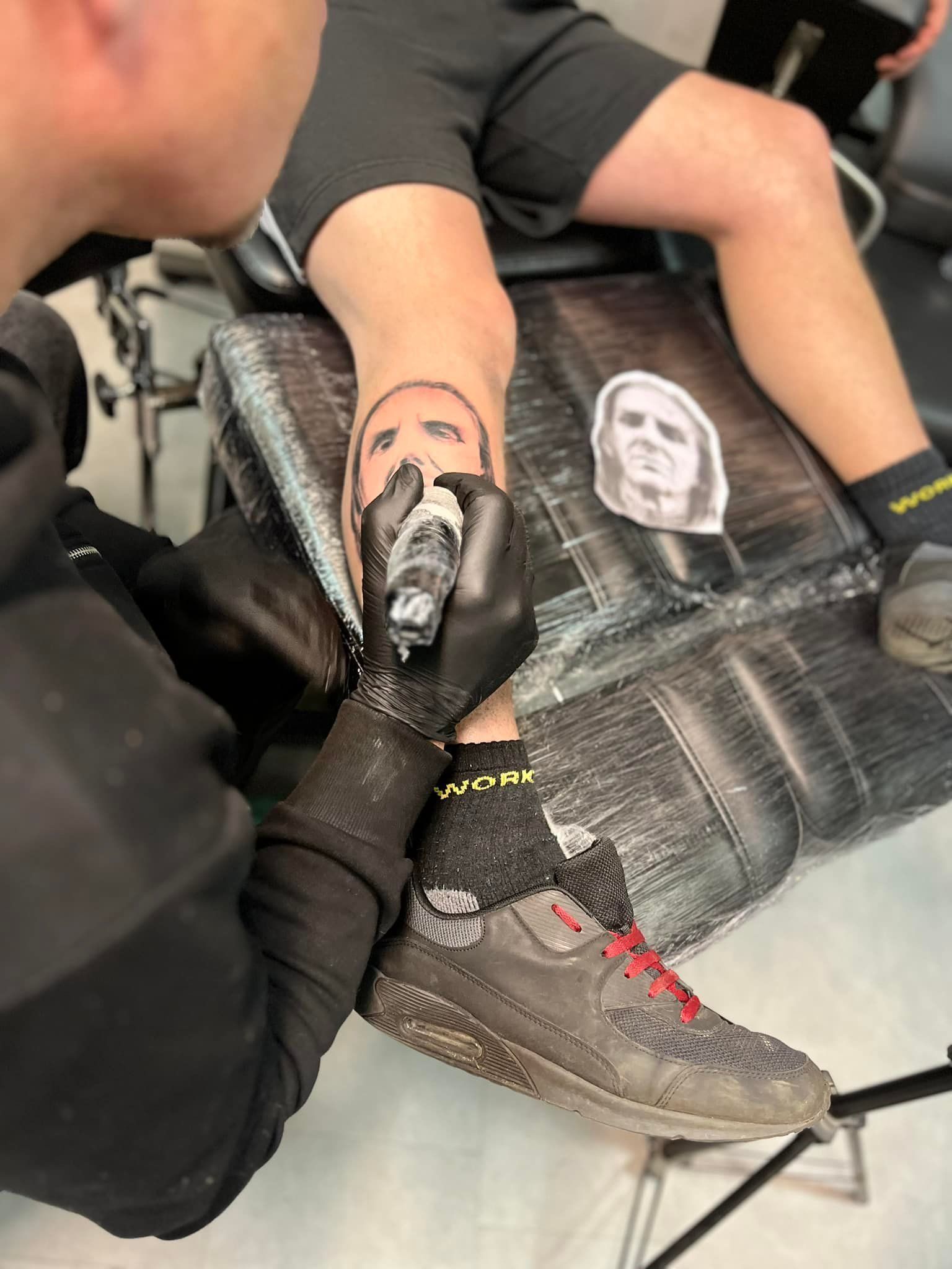 Persoon laat een gezichtstatoeage op zijn been zetten; kunstenaar aan het werk. Zwarte inkt, studio-omgeving.