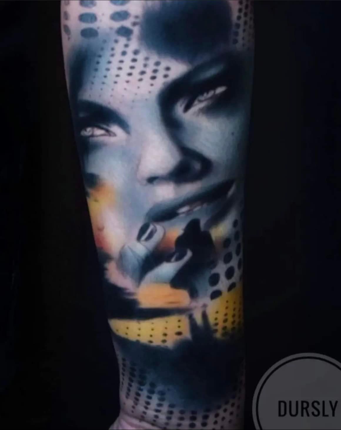 Tatoeage op een arm van een vrouwengezicht in de kleuren blauw, geel en zwart, abstract ontwerp met stippenpatroon.