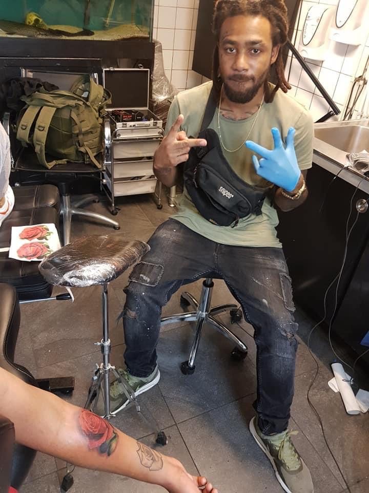 Man met dreadlocks en heuptasje maakt gebaren bij tatoeëerder. Tatoeage op arm in uitvoering. Tattoostudio.