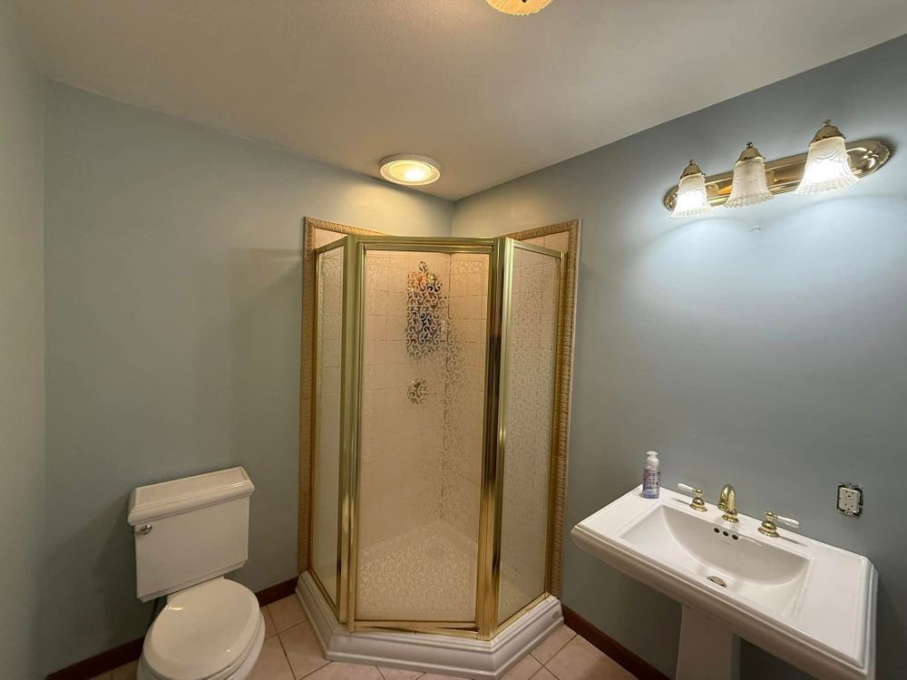 Bathroom with shower stall, toilet, and sink in soft blue walls and gold trim.