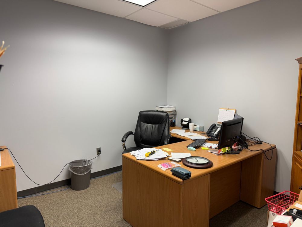 Empty office with a desk, black chair, computer equipment, and a trash can against gray walls.