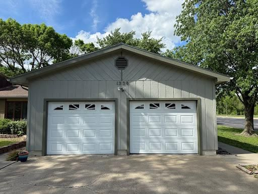 Two-car gray garage with white doors and a tree-lined driveway under a blue sky