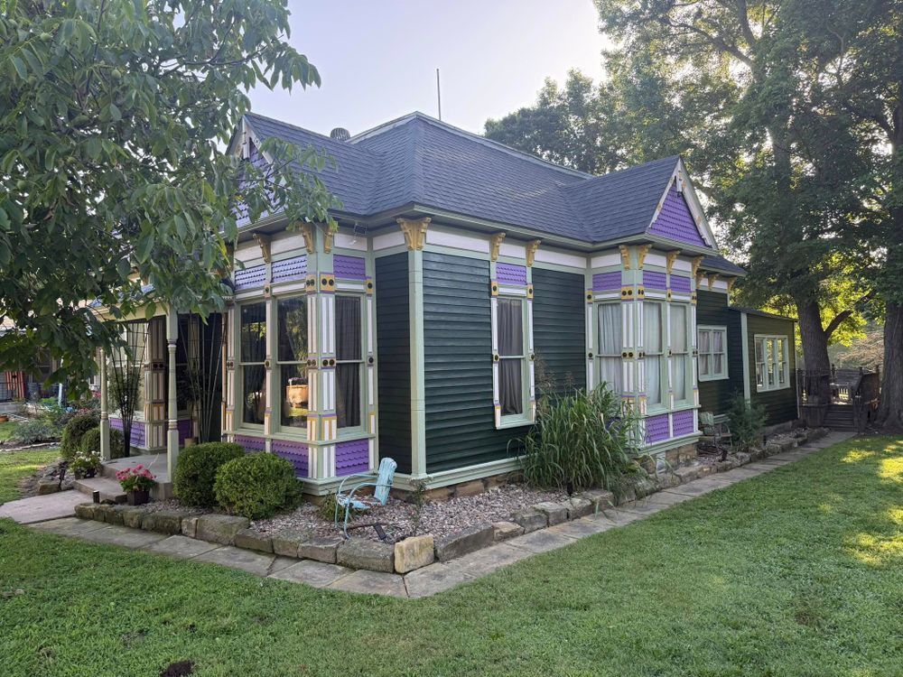 Small Victorian-style house with purple trim, dark green siding, and a front porch amid trees and lawn
