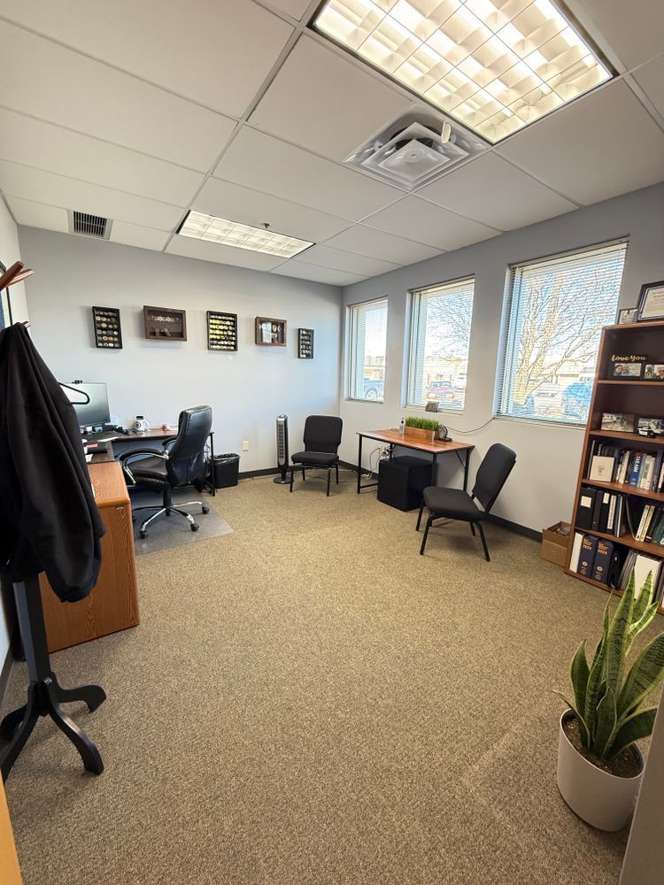 Small office with desks, chairs, bookshelves, windows, and potted plants under fluorescent lights.