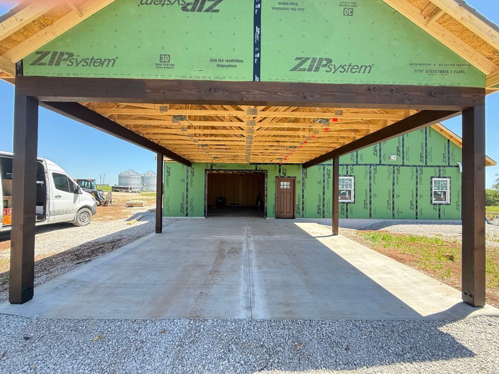 Covered carport with wooden roof framing and steel posts on a concrete slab beside a green house under construction