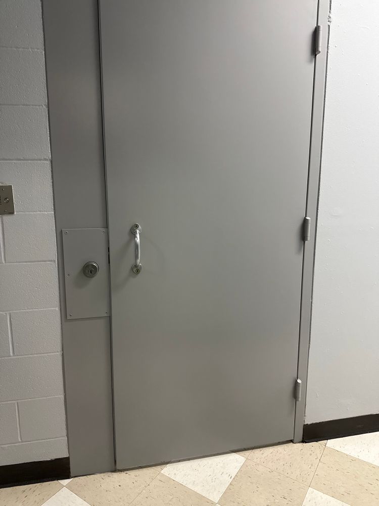 Gray metal door in a hallway with cinderblock wall and a white floor marker beneath it