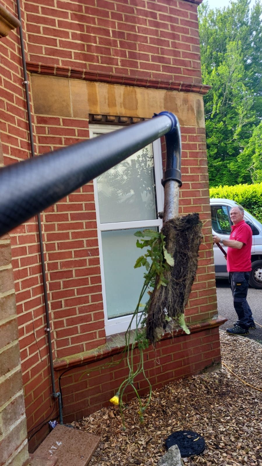 A man in a red shirt is standing in front of a brick building. the gutter cleaner is full of leaves and moss