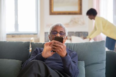 An elderly man is sitting on a couch using a cell phone.