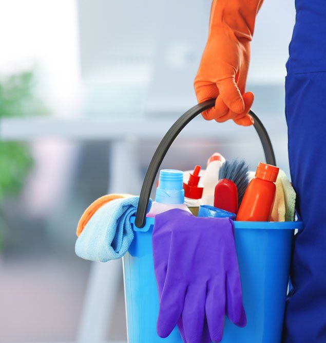 Person in orange gloves and blue scrubs holds a blue bucket of cleaning supplies, indoors.