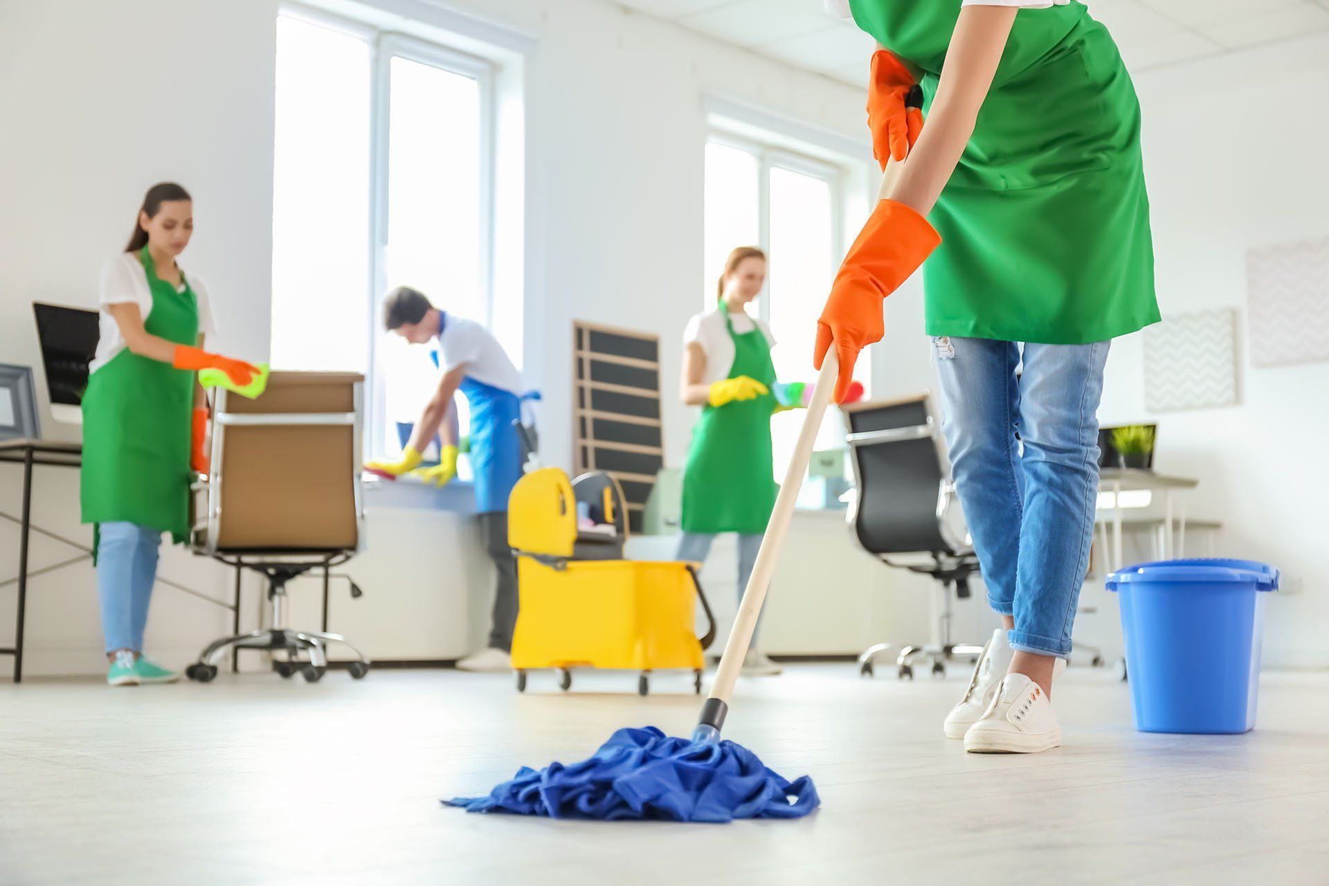 Office cleaners wearing green aprons and gloves cleaning an office, mopping floor, and wiping surfaces.