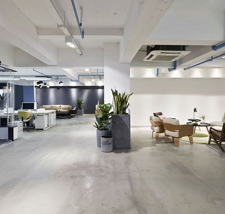 Modern office interior with desks, seating areas, plants, and exposed ceiling pipes; mostly white, gray, and blue tones.