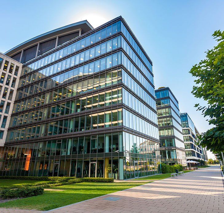Modern glass-walled office buildings with a blue sky background, trees, and walkway.