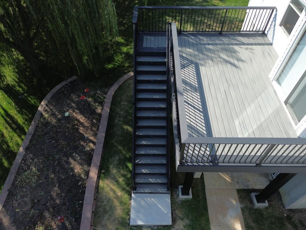 Overhead view of a deck with stairs. Dark metal railing, gray composite decking, and concrete pathway.