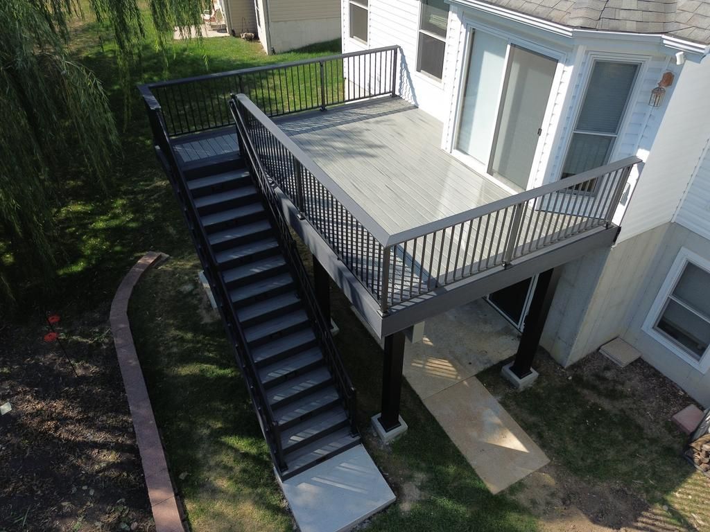 Elevated wooden deck with stairs and black railing attached to a two-story house.