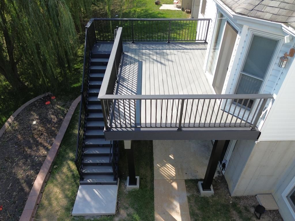 Elevated deck with stairs, supported by black pillars. Gray decking, black railing. Near a house and green lawn.
