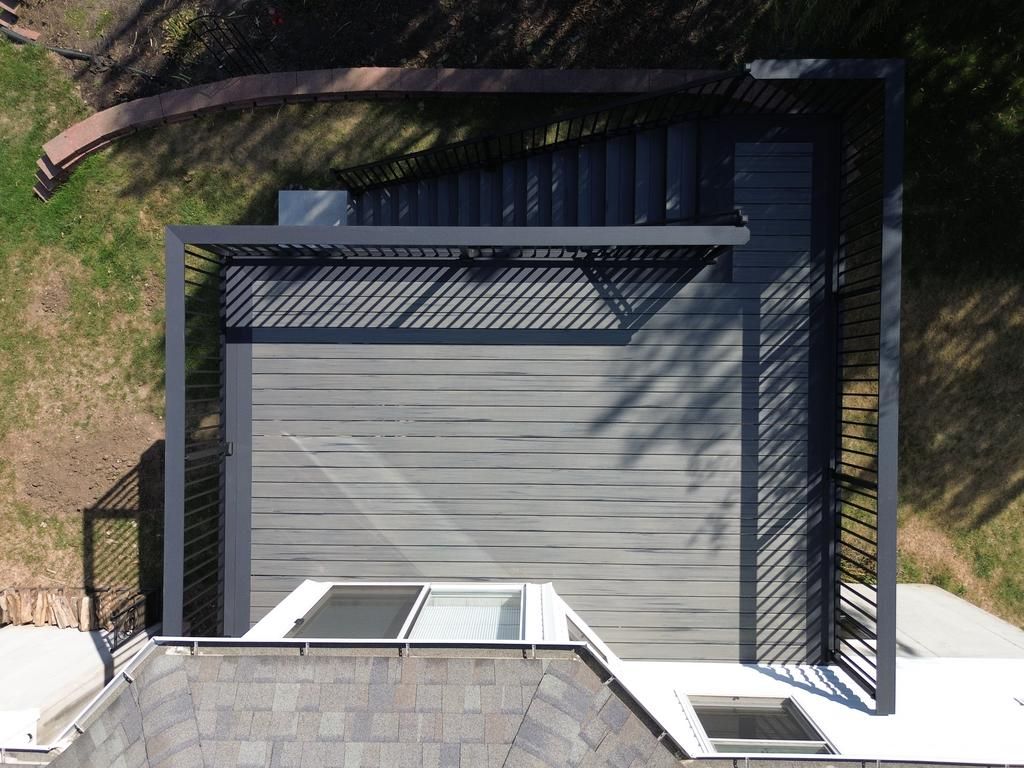 Overhead view of a modern gray deck with a pergola, steps, and house roof with skylights, set on a green lawn.