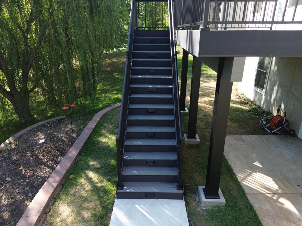 Outdoor staircase leading from a deck to a concrete landing, surrounded by grass and a weeping willow tree.