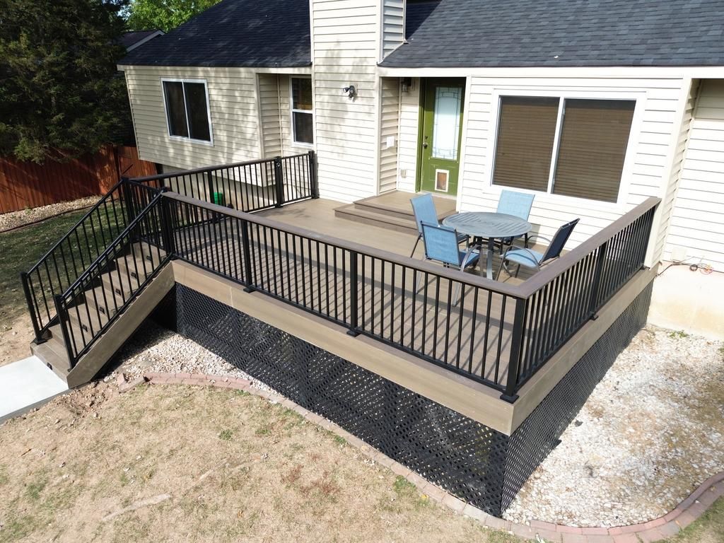 Deck with black railing, stairs, and table with chairs, attached to a house with black shutters and a green door.