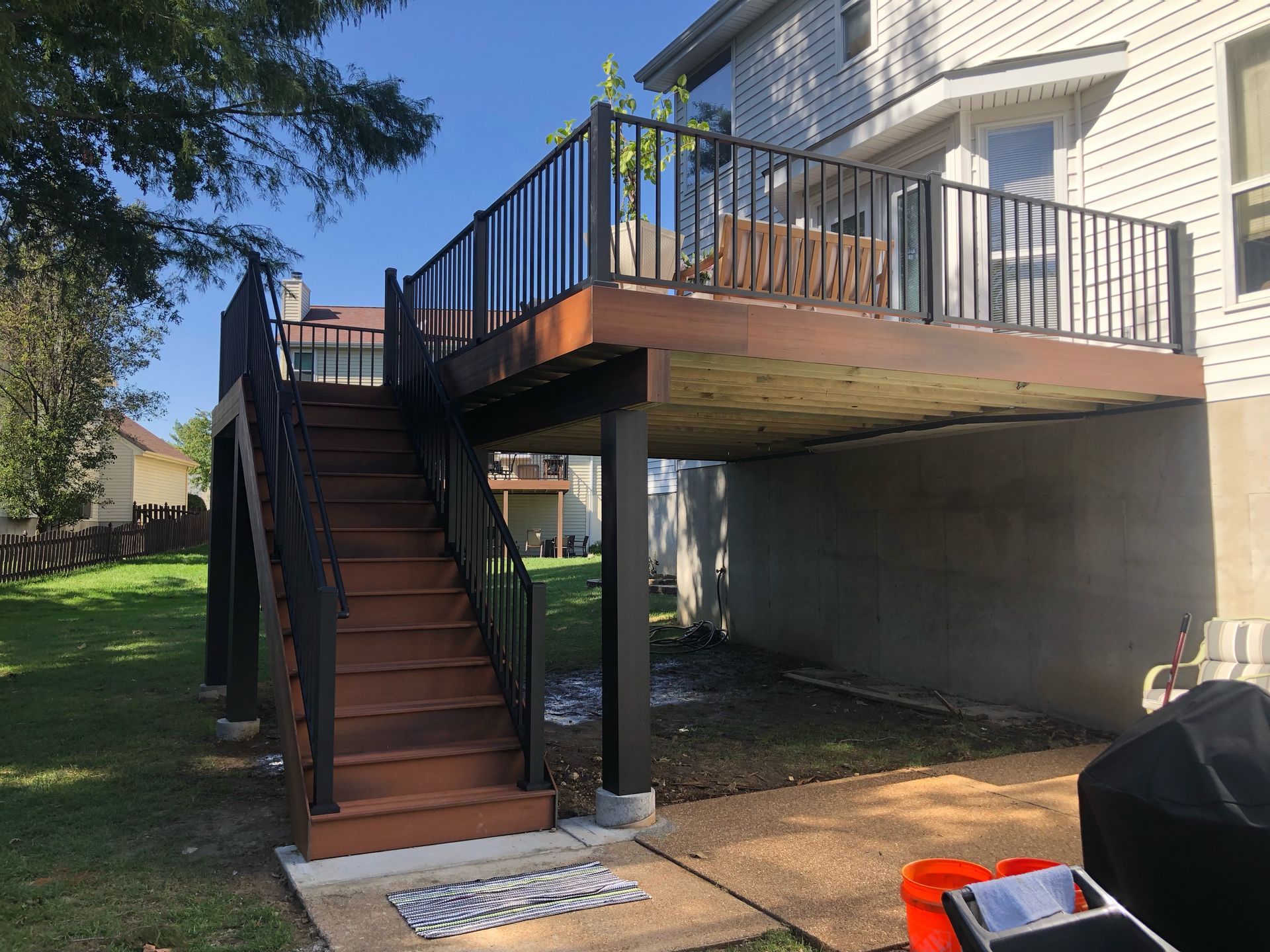 A two-story deck with stairs. Brown decking and stairs, black railings, and a gray house exterior. Sunny, outdoor setting.