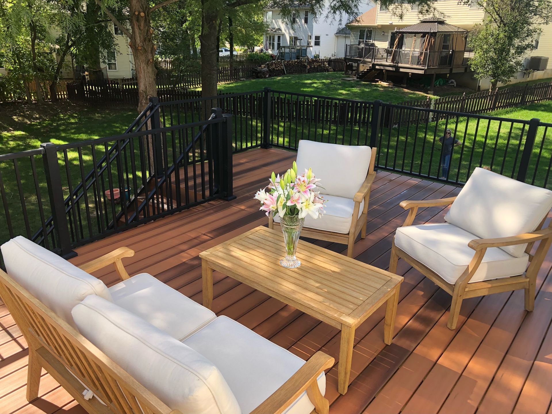 Outdoor deck with light wood furniture, white cushions, and flowers; surrounded by black railing and greenery.