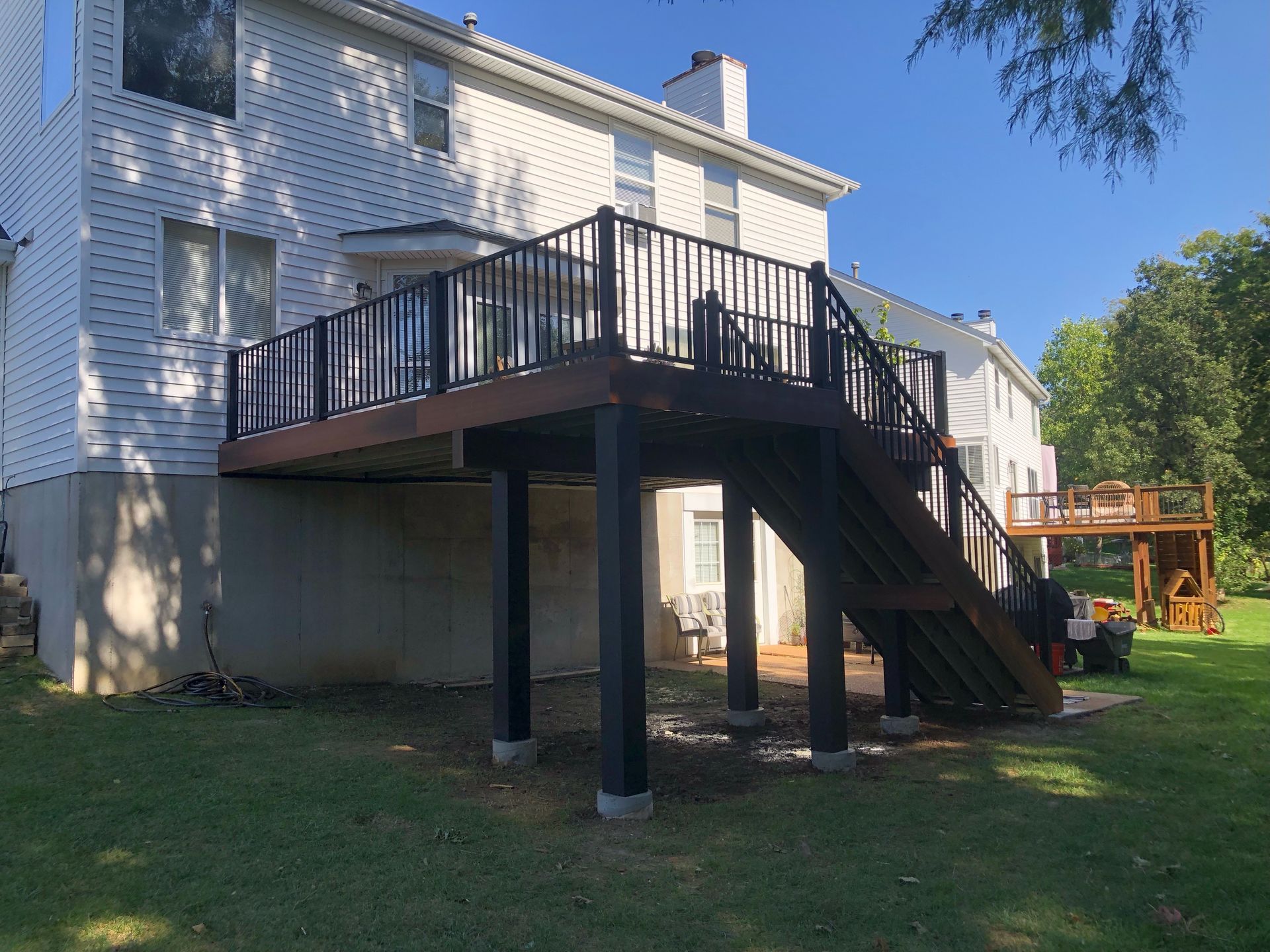 Two-story house with a wooden deck and stairs; dark brown railing and supports, green lawn.