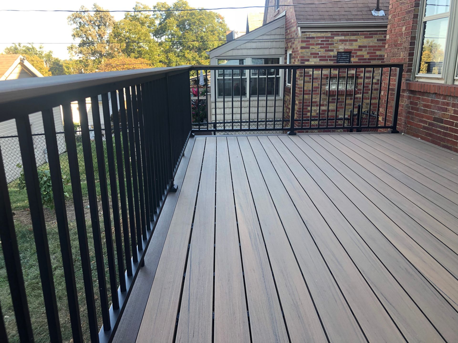 Black railing surrounds a composite deck. A brick house and trees are in the background.