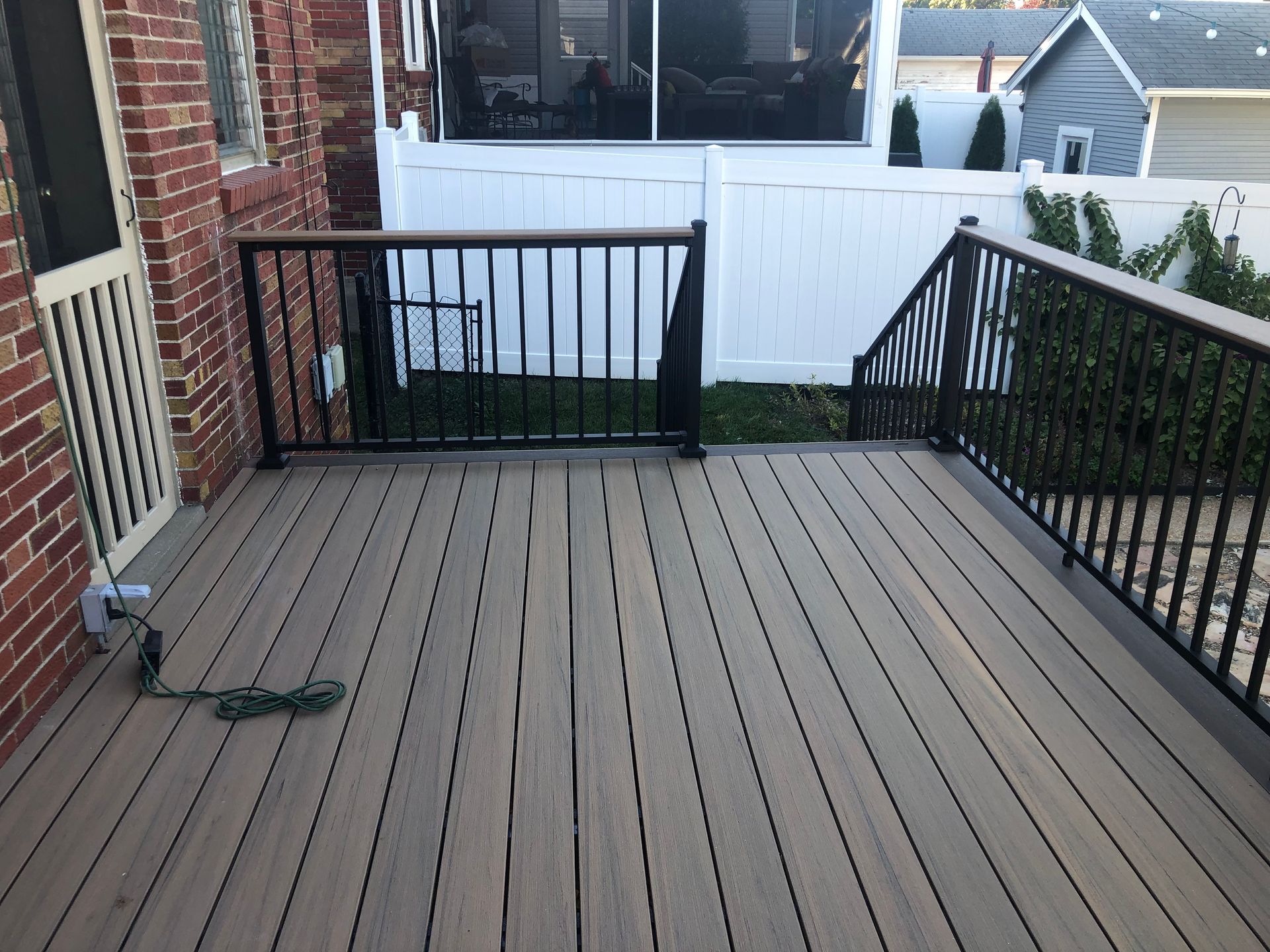 Wooden deck with black railings and steps leading to a white fence. Building in the background.