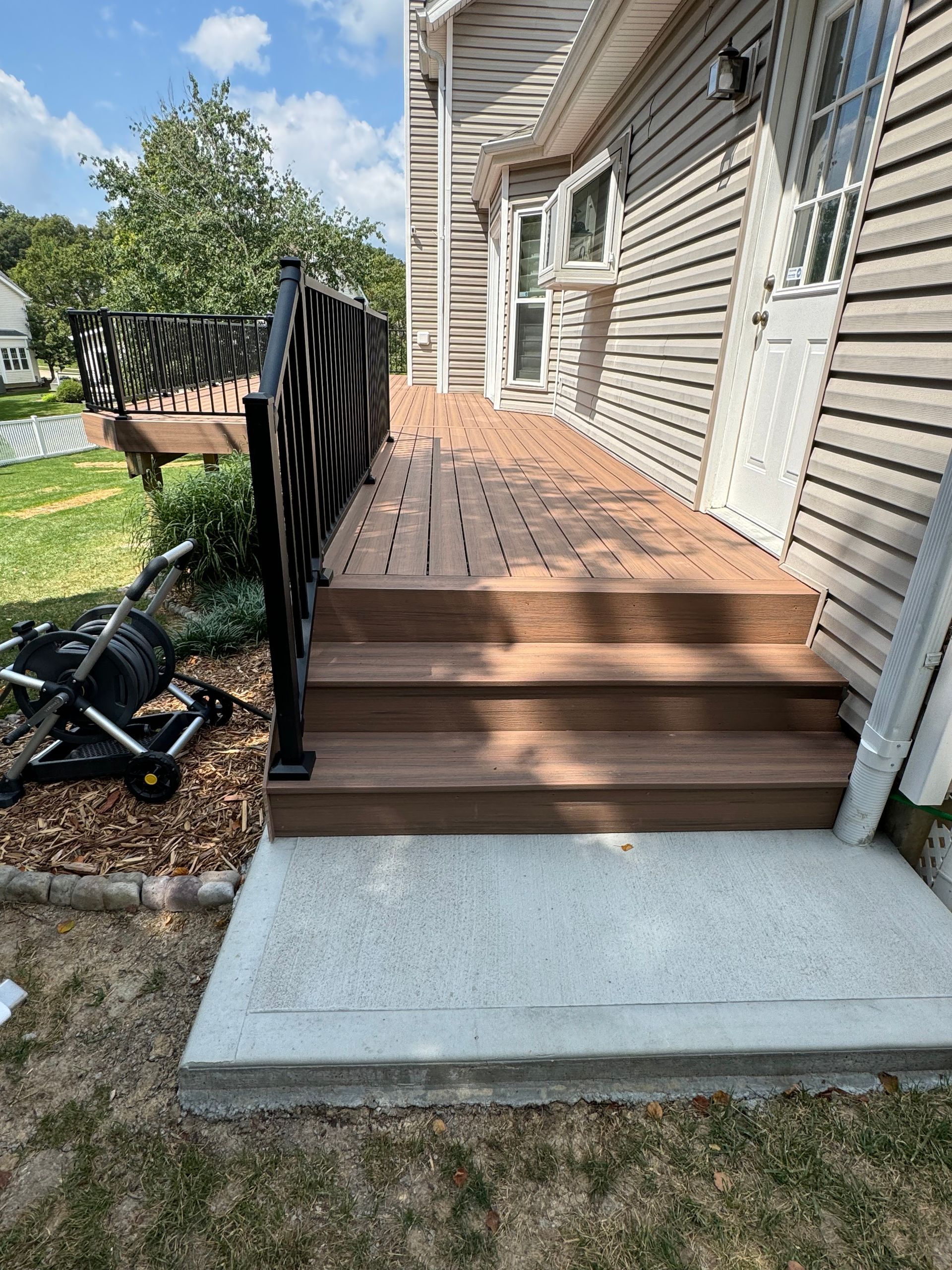 Wooden deck with stairs leading to a house. Black railing. Concrete pad at base.