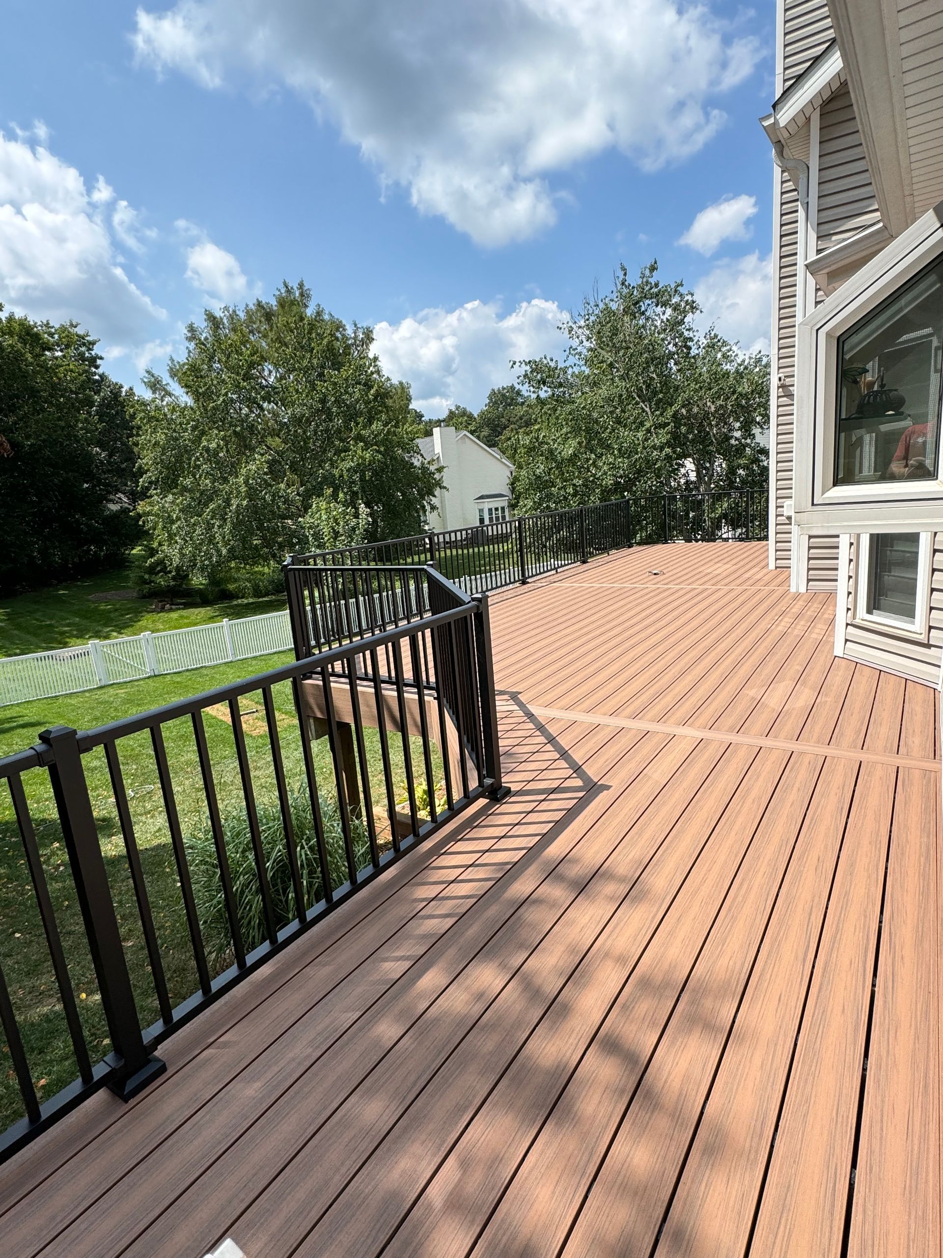 Composite deck with black railing, overlooking a green yard and trees under a blue sky.