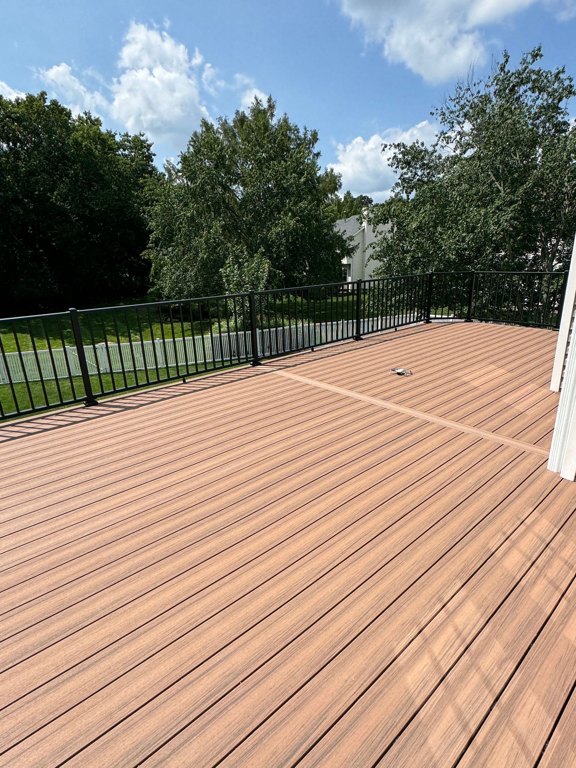 Brown composite deck with black railing overlooking a green lawn and trees under a blue sky.