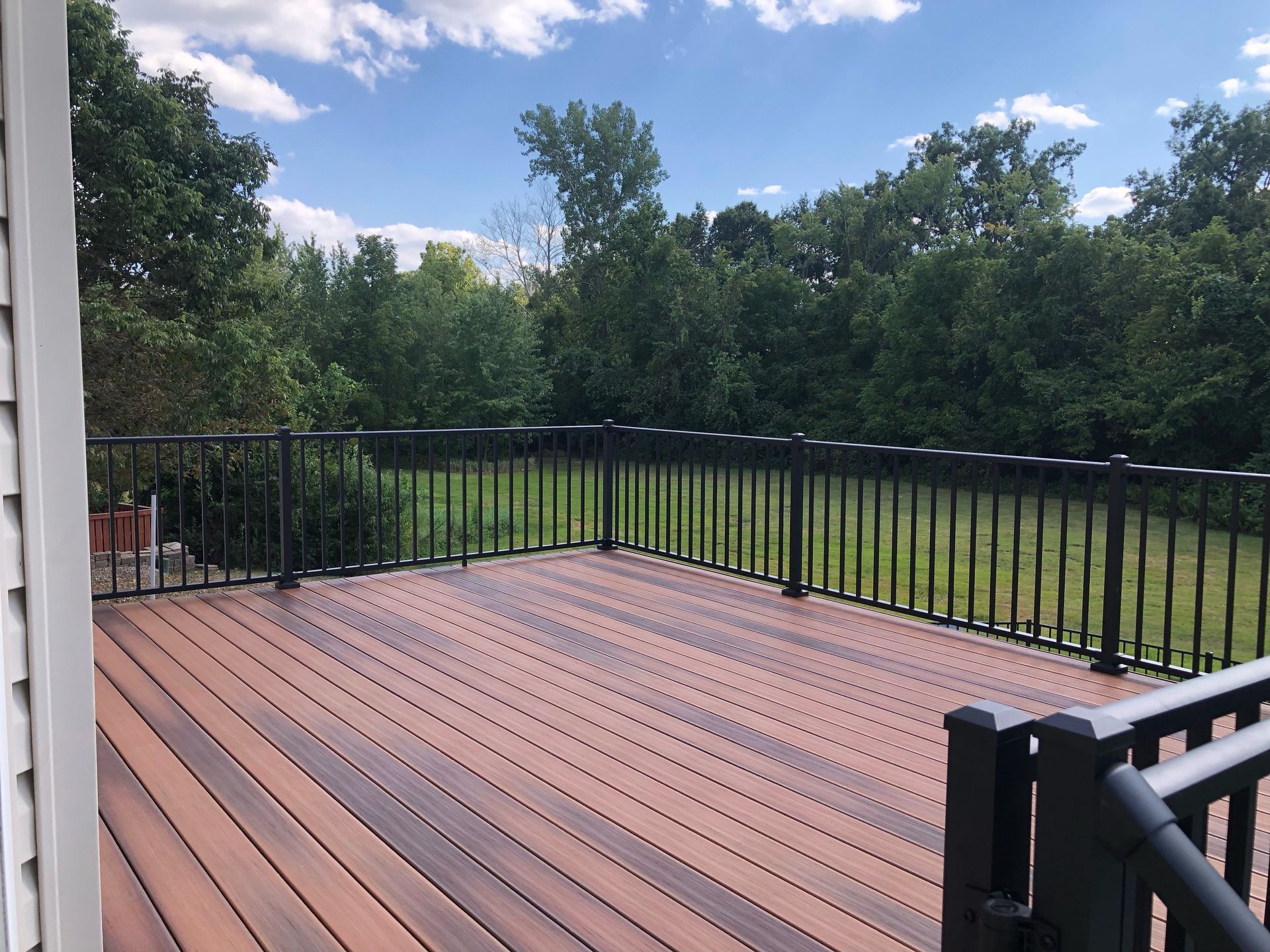 Wooden deck with black railing overlooking a green yard and trees under a blue sky.