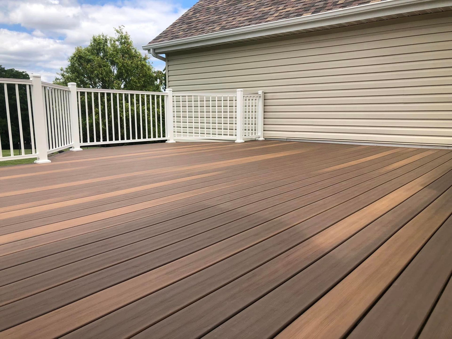 Wooden deck with white railing next to a house with tan siding under a blue sky.