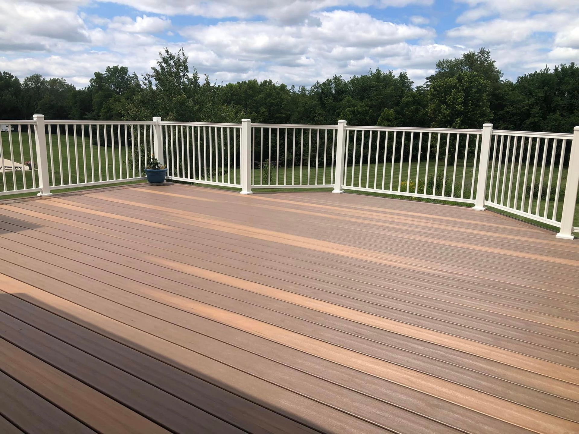 Brown composite deck with white railing, overlooking a green treeline under a partly cloudy sky.