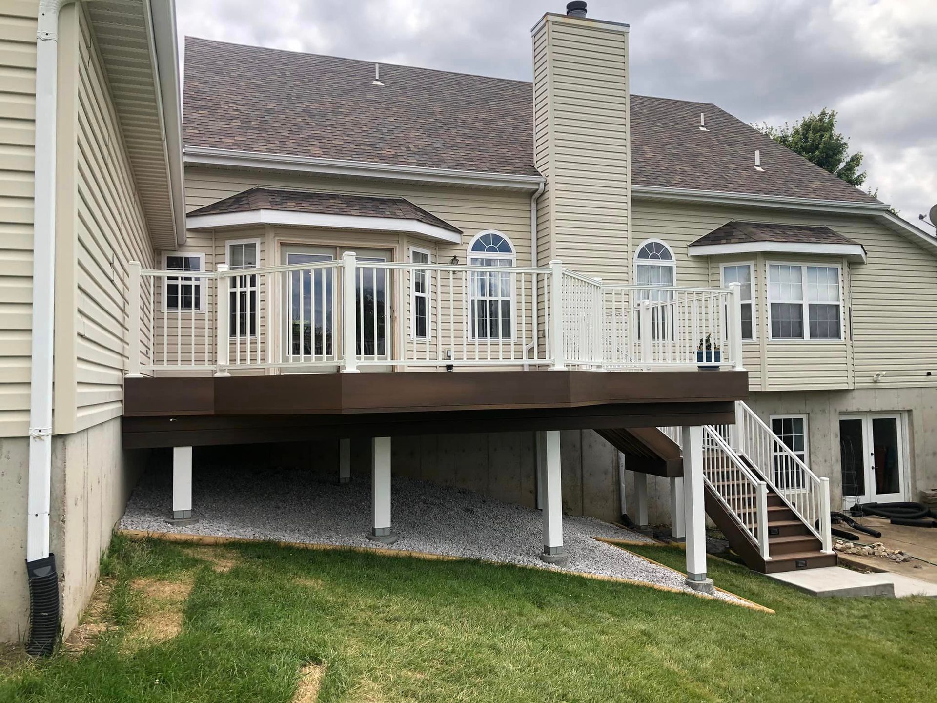 Back of a house with a brown deck, white railing, and stairs. Deck is over a gravel bed.