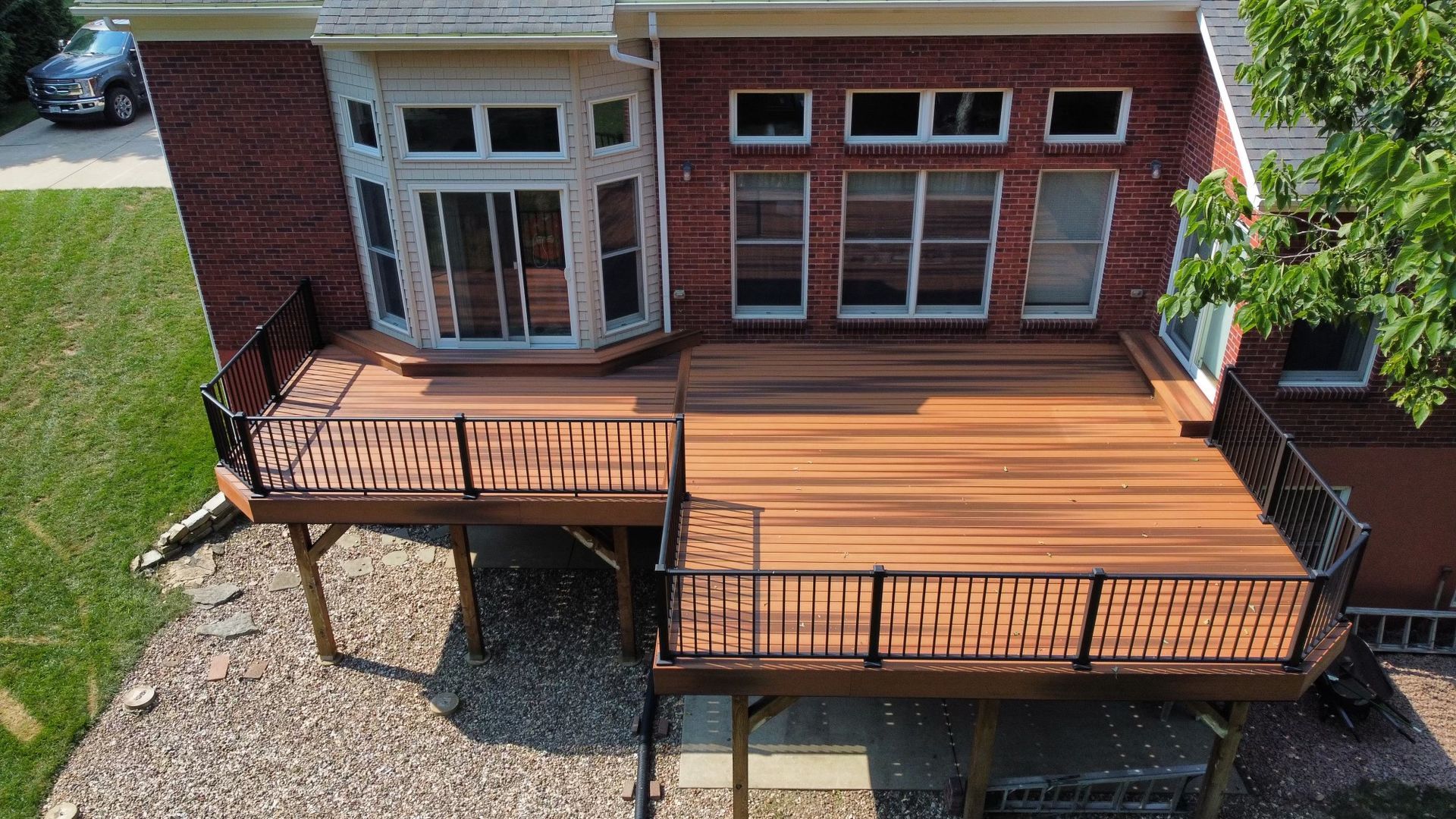 Elevated wooden deck attached to a brick house. Black railings, brown deck boards, and a backyard setting.