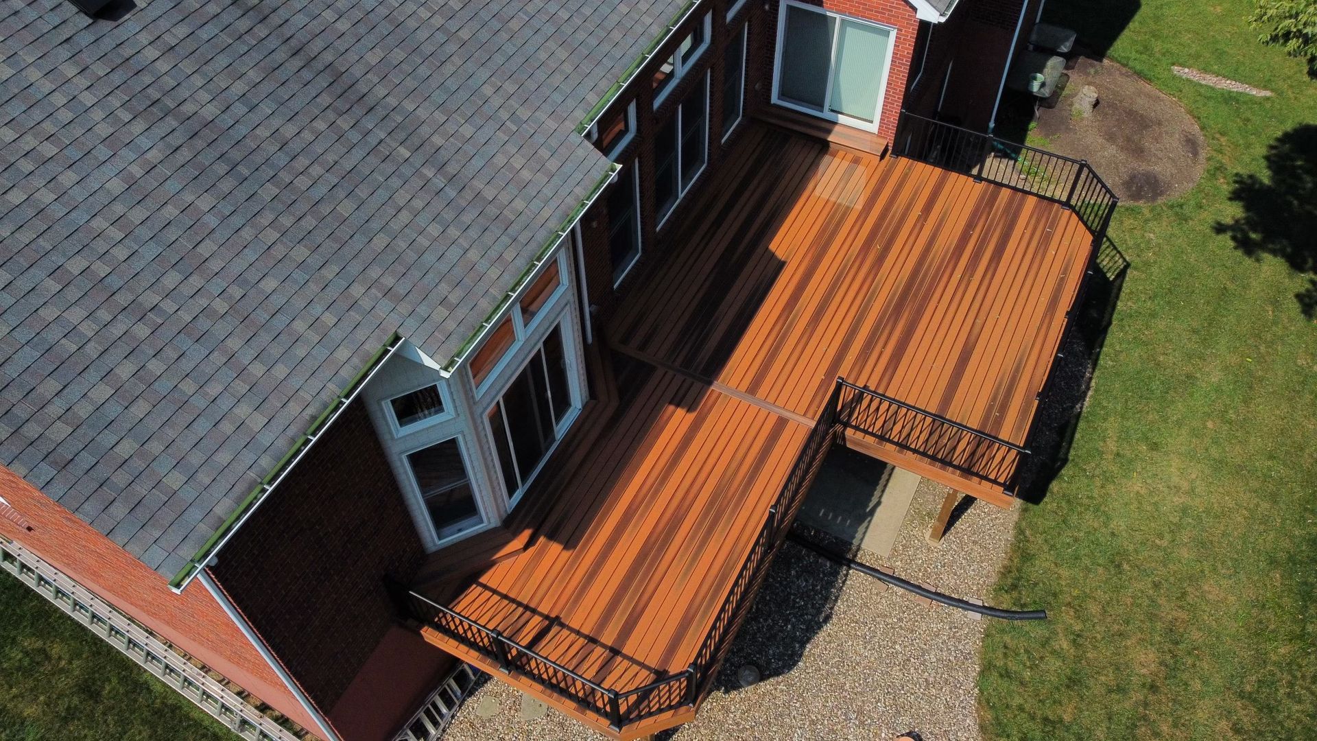 Aerial view of a wooden deck attached to a two-story house with a black railing, next to green grass.