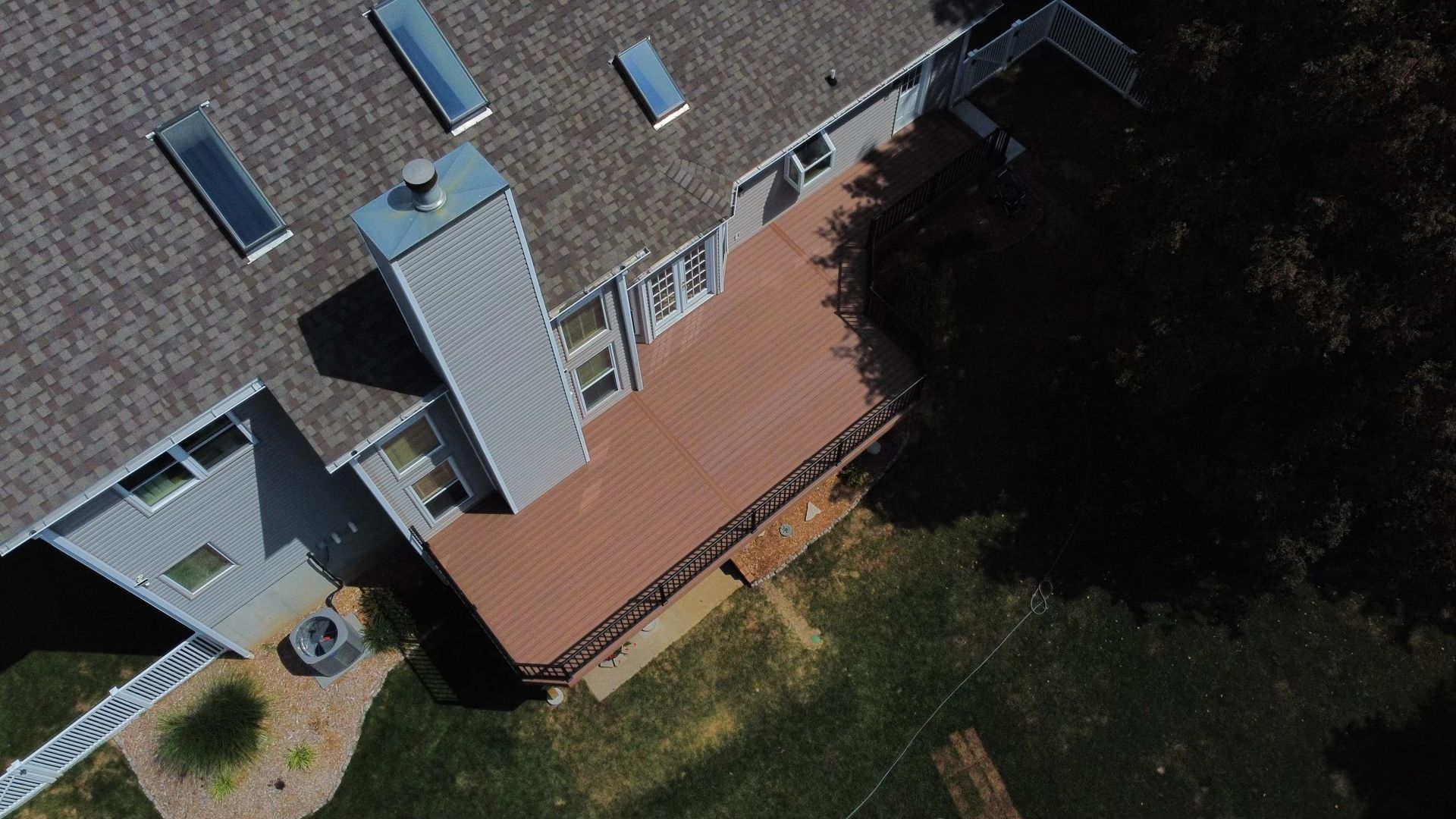 Overhead view of a house with a brown deck, gray roof, chimney, and surrounding green lawn.