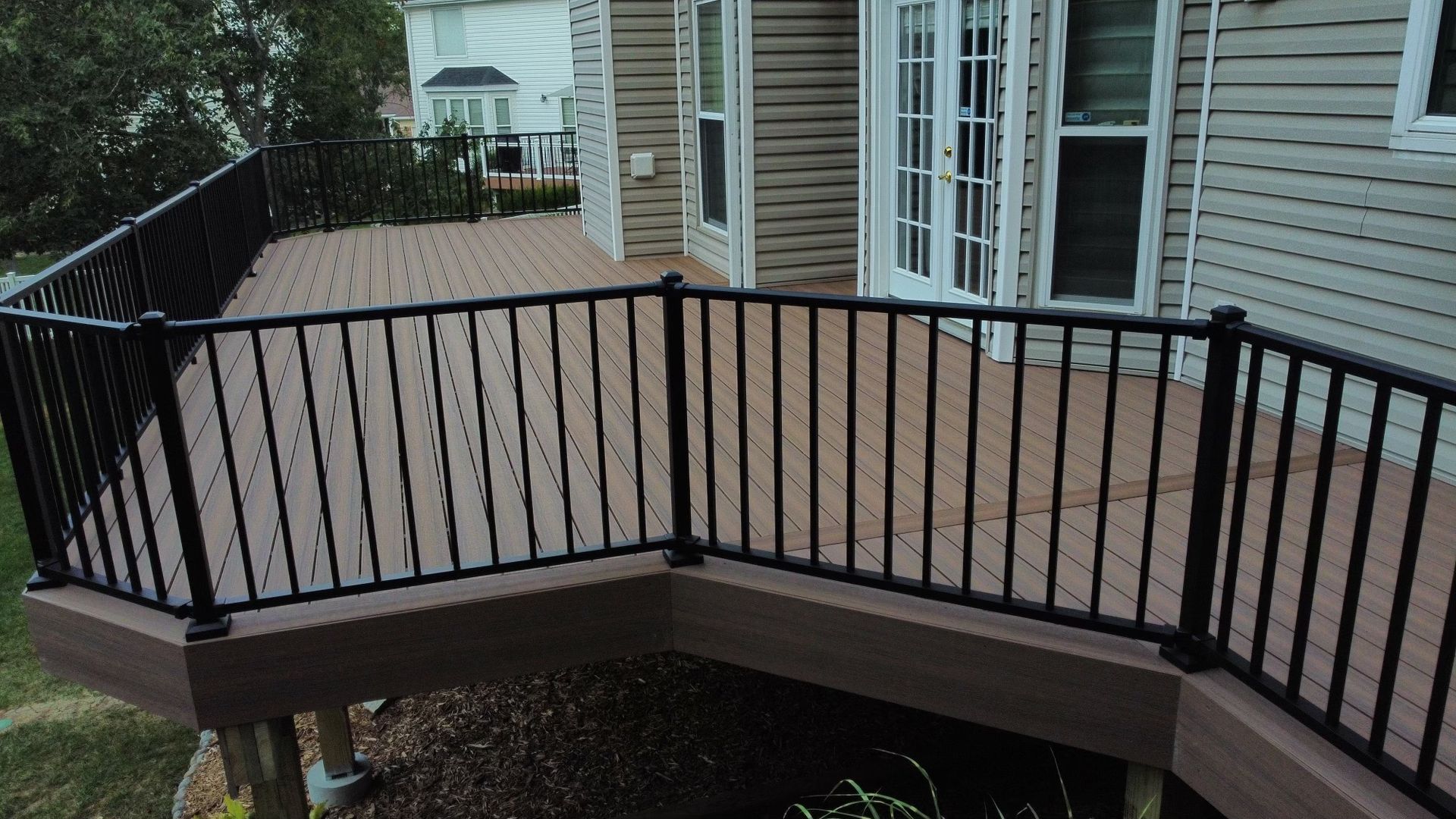 Brown composite deck with black metal railing, adjacent to a house with white doors and siding.