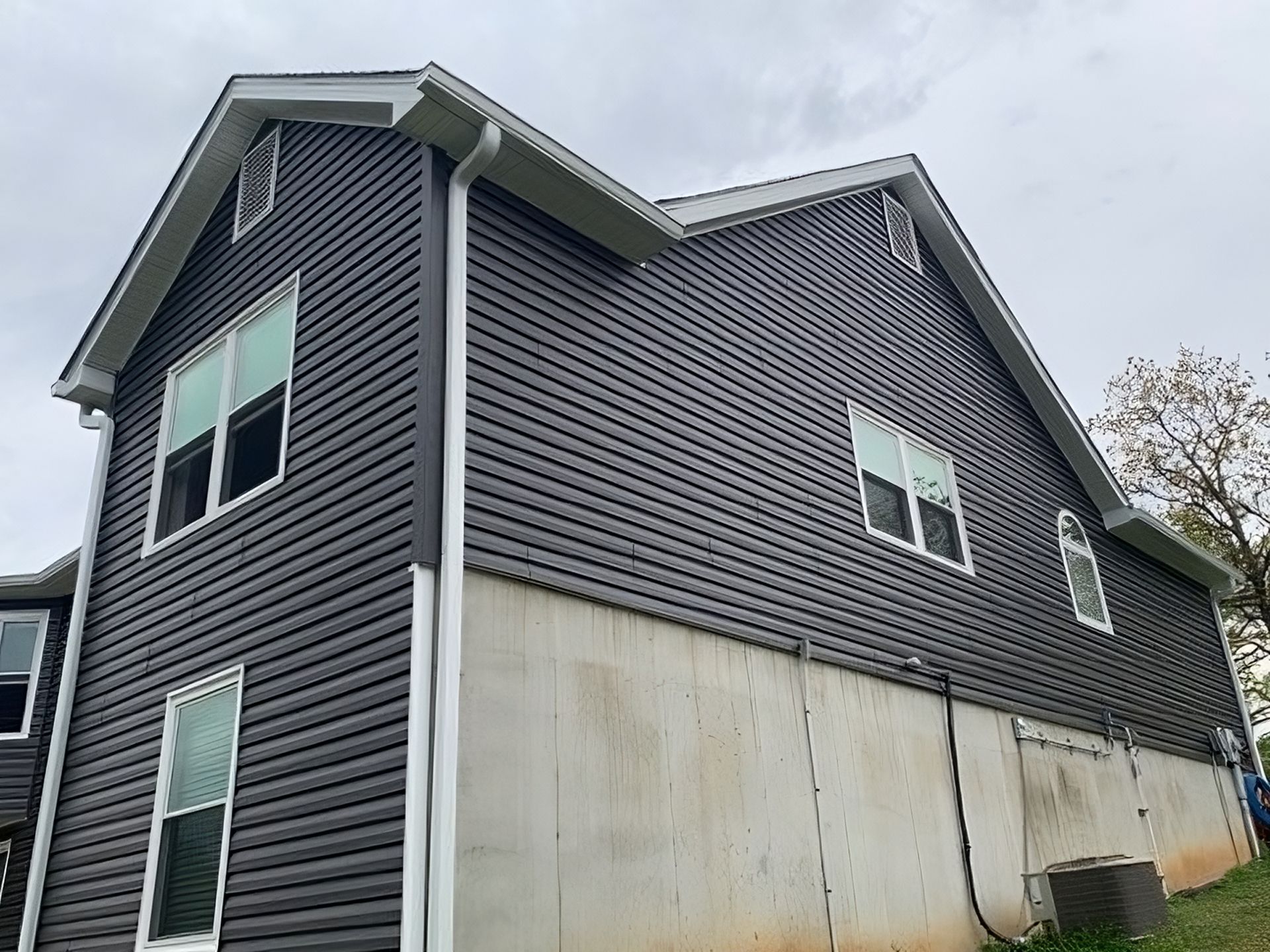 Dark gray siding on a two-story house with white trim. Concrete foundation below. Overcast sky.